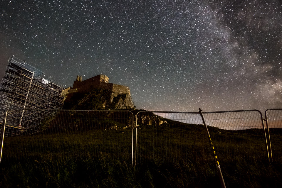 Lindisfarne Castle on Holy Island at night with the Milky Way galaxy visible in the starry sky above, scaffolding covering part of the historic building and security fencing in the foreground