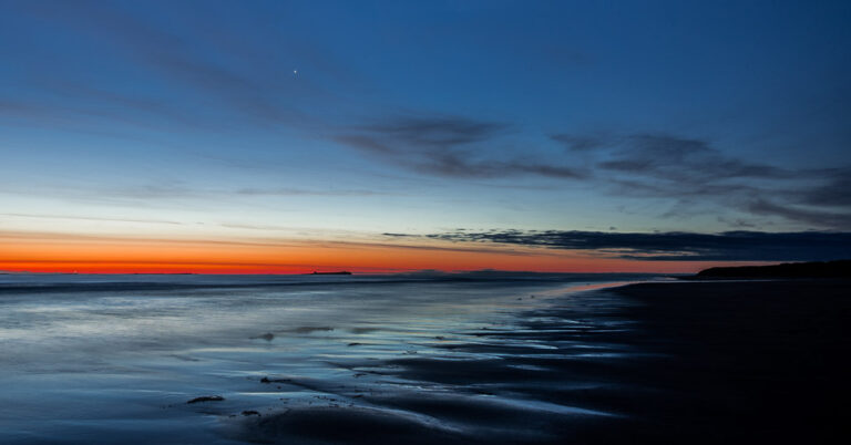 Blue hour sunset at Bamburgh Beach with orange and blue gradient sky reflected in wet sand, Farne Islands visible as dark silhouettes on the horizon