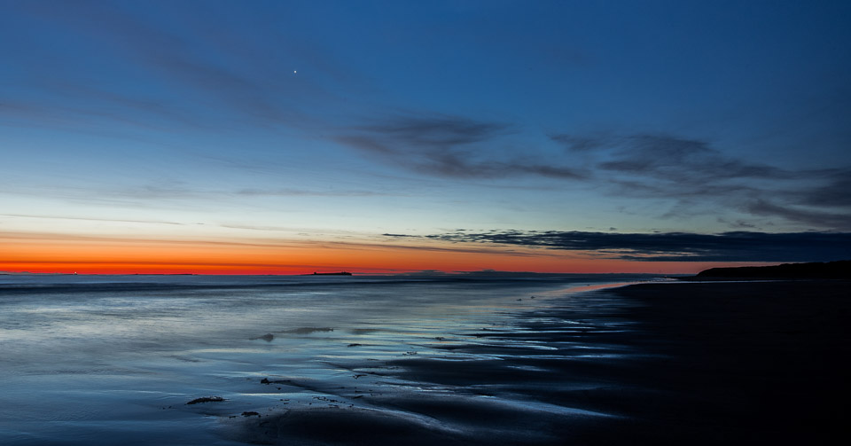 Blue hour sunset at Bamburgh Beach with orange and blue gradient sky reflected in wet sand, Farne Islands visible as dark silhouettes on the horizon