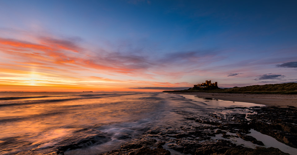 Dawn at Bamburgh Beach with dramatic orange and pink sunrise sky reflected in wet sand and gentle waves, Bamburgh Castle silhouetted on rocky outcrop with sunlight illuminating its walls