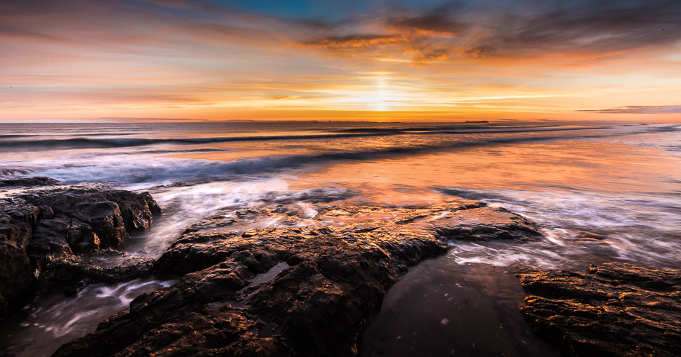 Dawn sunrise over the North Sea at Bamburgh Beach with dramatic orange and pink sky reflected in water, rocky outcrops in foreground and smooth wave motion captured with long exposure