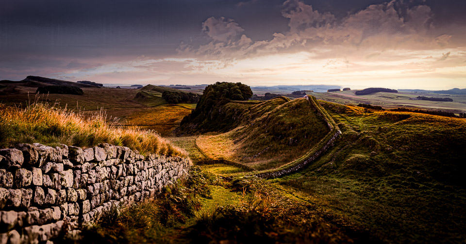 Early morning view of Hadrian's Wall at Whin Sill with ancient stone wall in foreground, undulating landscape creating deep shadows in low light, and dramatic sky with storm clouds overhead