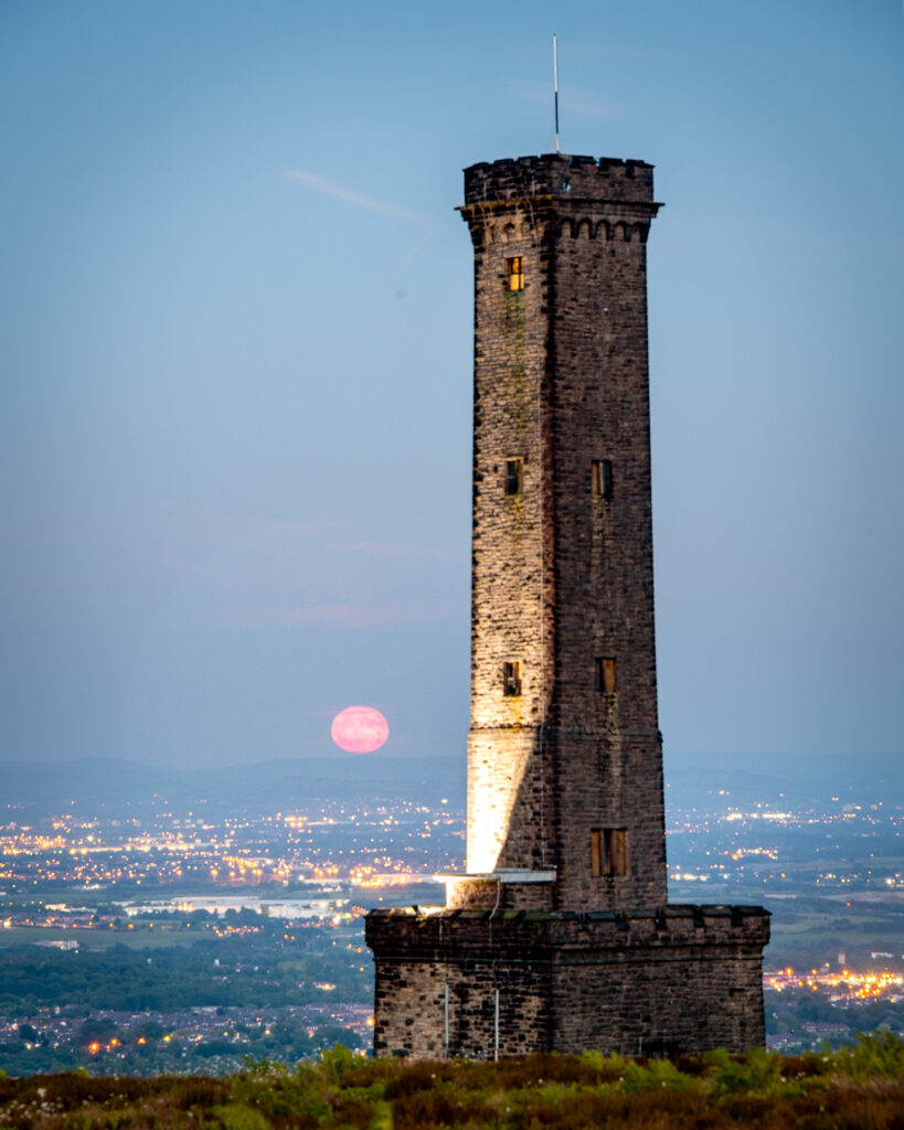 Peel Monument tower at dusk with pink full moon rising behind it and illuminated Manchester suburbs sprawling across the valley below