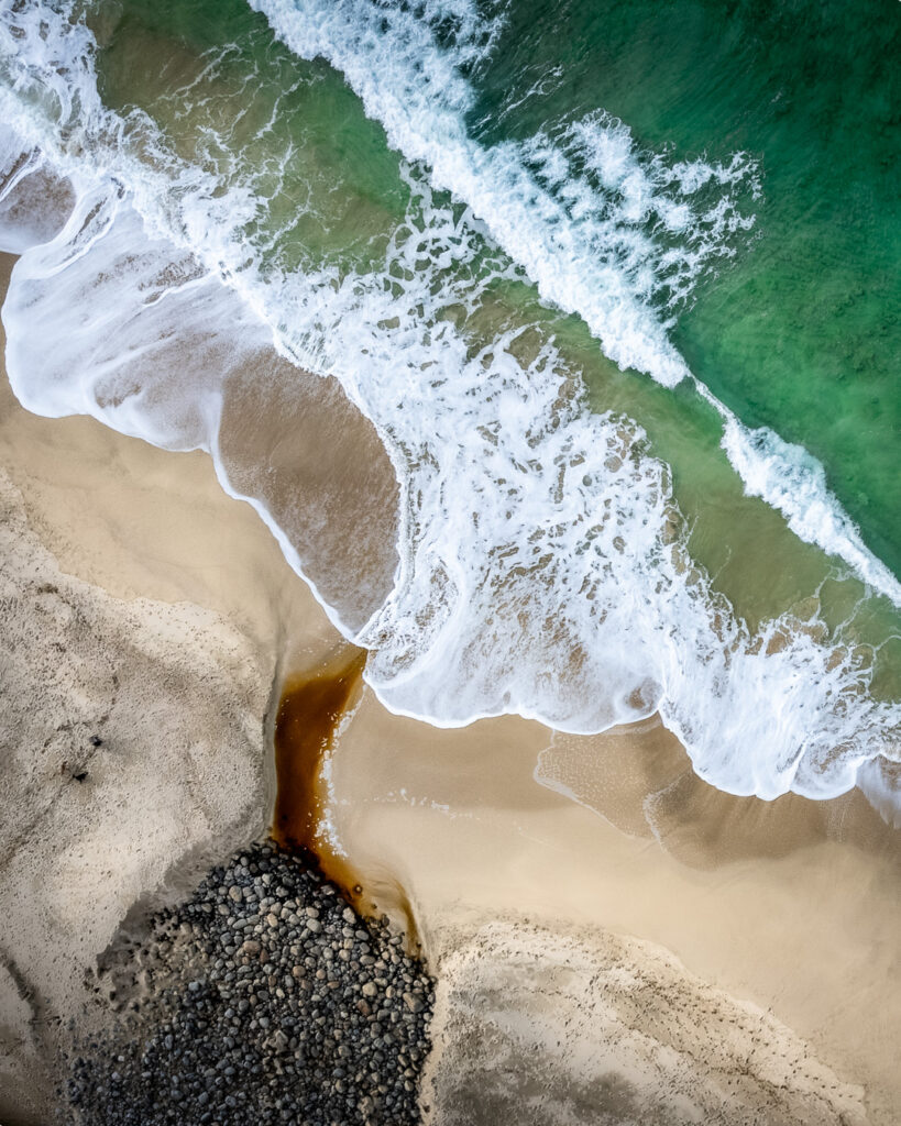 Drone view of Dail Mor beach, with a peat-stained Highland burn winding across golden sand into emerald Atlantic waters, white waves breaking along the shore, and a cluster of dark rounded stones.