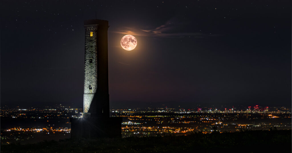 Silhouetted Peel Monument tower against night sky with bright full moon overhead and Manchester city lights sprawling across the horizon