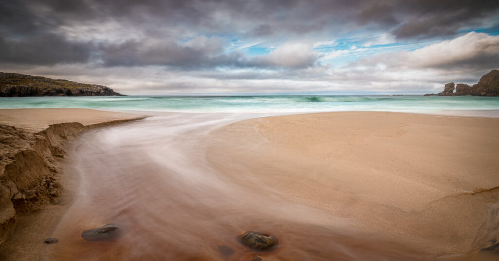 Long exposure of Dail Mòr beach, Isle of Harris, with a stream winding over golden sand into turquoise Atlantic waves beneath brooding, cloud-filled skies and rugged headlands.