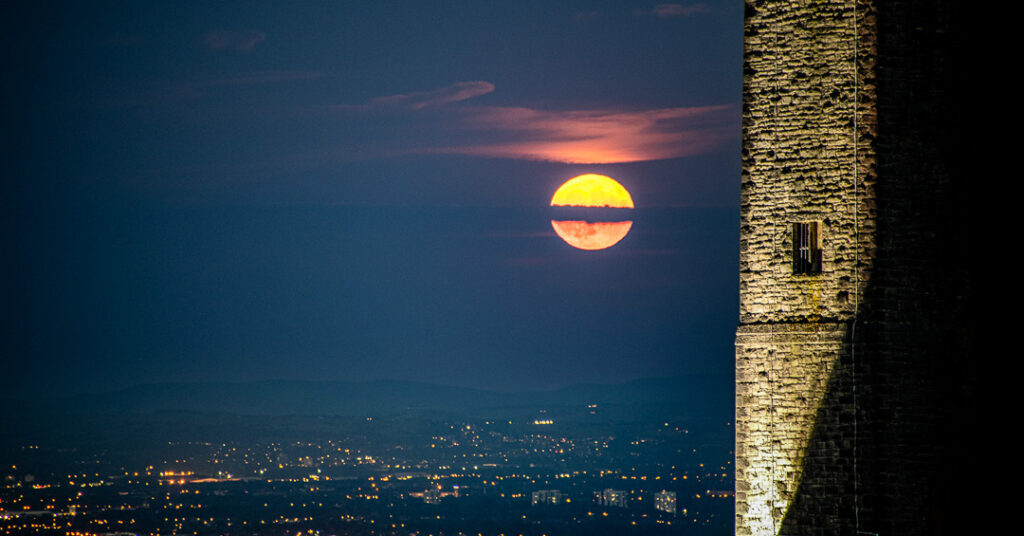 Full moon rising through dramatic sky with floodlit stone wall of Peel Monument in foreground and Manchester city lights twinkling in valley below