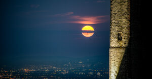 Full moon rising through dramatic sky with floodlit stone wall of Peel Monument in foreground and Manchester city lights twinkling in valley below