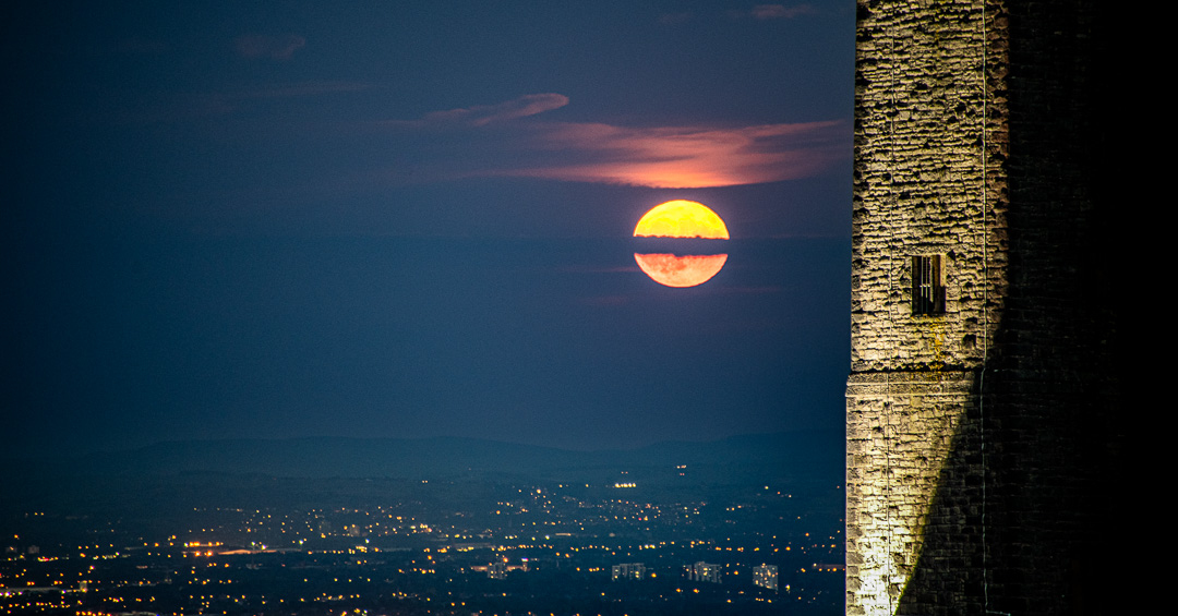 Full moon rising through dramatic sky with floodlit stone wall of Peel Monument in foreground and Manchester city lights twinkling in valley below