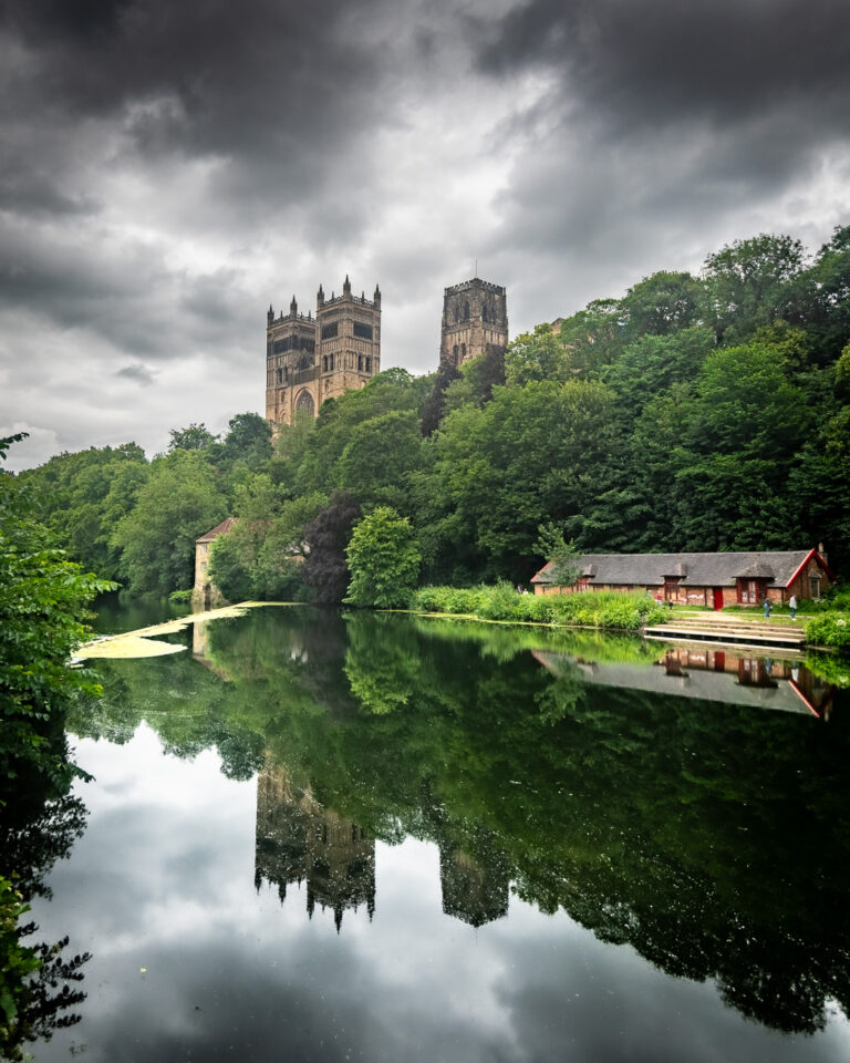 Durham Cathedral with twin towers rising above lush green trees, reflected in the calm waters of River Wear below, under dramatic cloudy sky by Michael Dutson Landscape Photography